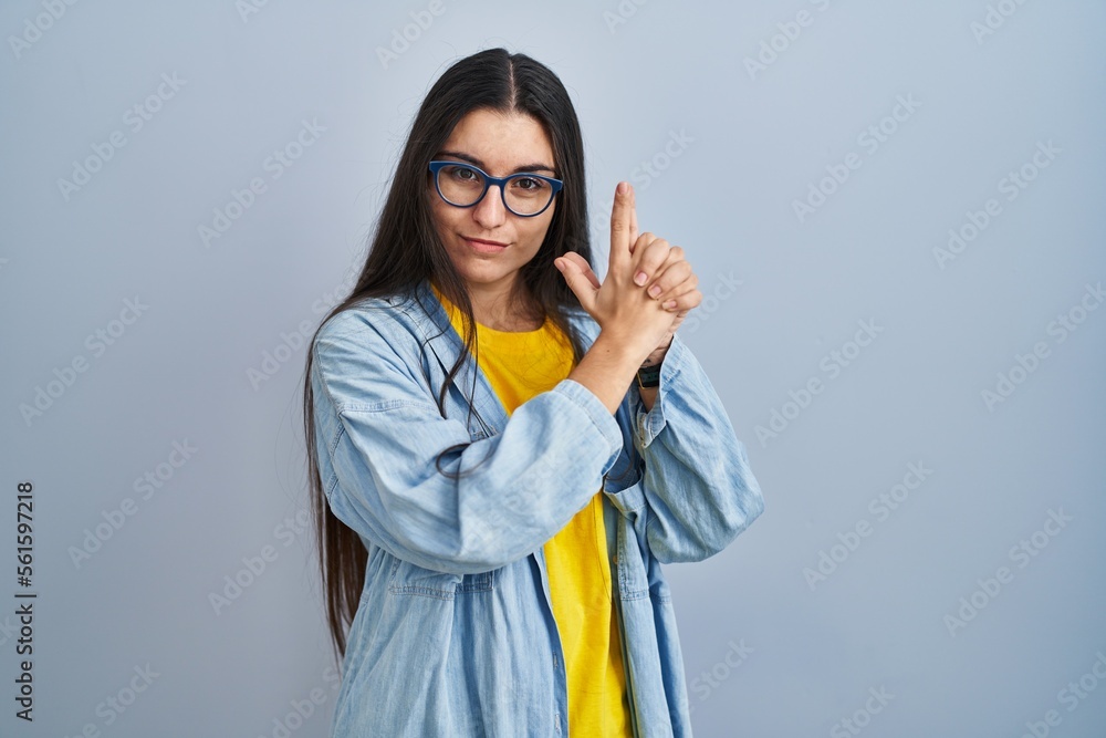 Young hispanic woman standing over blue background holding symbolic gun with hand gesture, playing killing shooting weapons, angry face