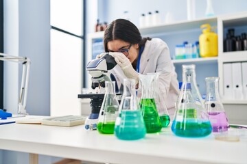 Poster - Young hispanic woman wearing scientist uniform using microscope at laboratory