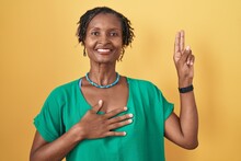 African Woman With Dreadlocks Standing Over Yellow Background Smiling Swearing With Hand On Chest And Fingers Up, Making A Loyalty Promise Oath