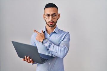 Poster - Young hispanic man working using computer laptop pointing with hand finger to the side showing advertisement, serious and calm face