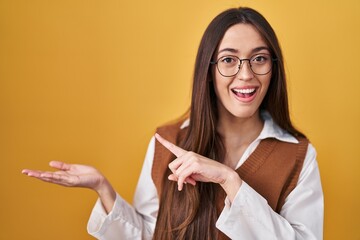 Canvas Print - Young brunette woman standing over yellow background wearing glasses amazed and smiling to the camera while presenting with hand and pointing with finger.