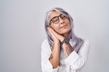 Wall Mural - Middle age woman with tattoos wearing glasses standing over white background sleeping tired dreaming and posing with hands together while smiling with closed eyes.