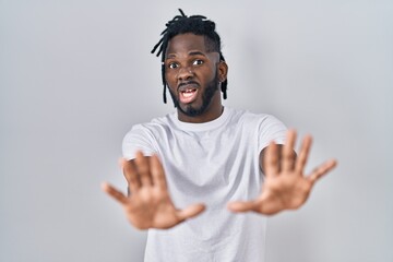 Poster - African man with dreadlocks wearing casual t shirt over white background afraid and terrified with fear expression stop gesture with hands, shouting in shock. panic concept.