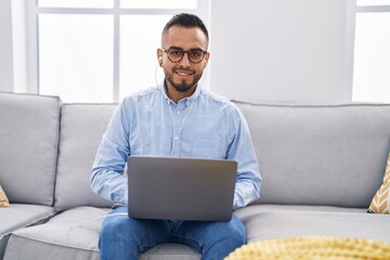 Sticker - Young hispanic man using laptop and earphones sitting on sofa at home