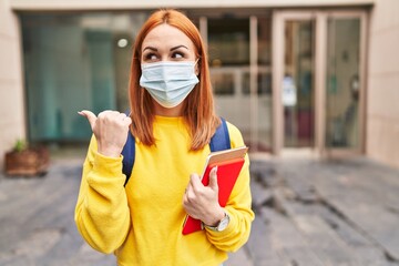 Sticker - Young woman wearing safety mask and student backpack holding books pointing thumb up to the side smiling happy with open mouth