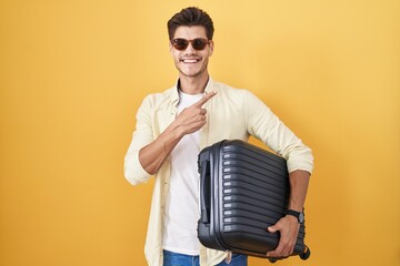 Canvas Print - Young hispanic man holding suitcase going on summer vacation cheerful with a smile on face pointing with hand and finger up to the side with happy and natural expression