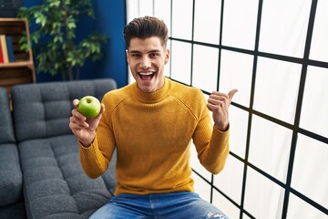 Wall Mural - Young hispanic man holding green apple pointing thumb up to the side smiling happy with open mouth