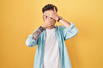 Wall Mural - Young hispanic man with tattoos standing over yellow background covering eyes and mouth with hands, surprised and shocked. hiding emotion