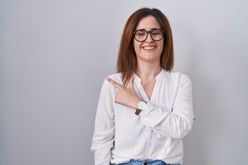 Sticker - Brunette woman standing over white isolated background cheerful with a smile on face pointing with hand and finger up to the side with happy and natural expression