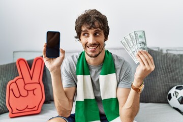 Wall Mural - Young hispanic man holding smartphone and dollars at home