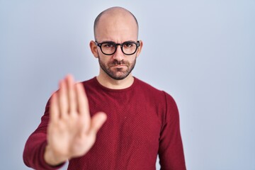 Wall Mural - Young bald man with beard standing over white background wearing glasses doing stop sing with palm of the hand. warning expression with negative and serious gesture on the face.