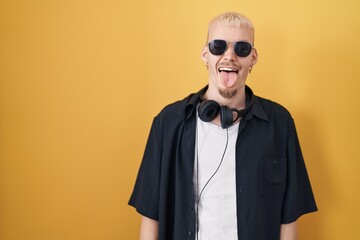 Sticker - Young caucasian man wearing sunglasses standing over yellow background sticking tongue out happy with funny expression. emotion concept.