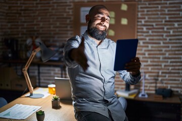 Wall Mural - Young hispanic man with beard and tattoos working at the office at night smiling friendly offering handshake as greeting and welcoming. successful business.