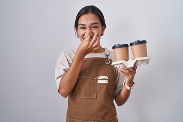 Wall Mural - Young hispanic woman wearing professional waitress apron holding coffee smelling something stinky and disgusting, intolerable smell, holding breath with fingers on nose. bad smell