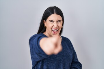 Poster - Young brunette woman standing over isolated background laughing at you, pointing finger to the camera with hand over body, shame expression