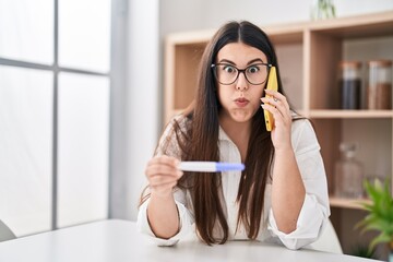 Canvas Print - Young brunette woman holding pregnancy test result speaking on the phone puffing cheeks with funny face. mouth inflated with air, catching air.