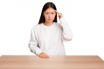 Wall Mural - Young asian woman sitting on a table isolated pointing temple with finger, thinking, focused on a task.