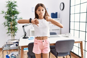 young hispanic girl standing at pediatrician clinic doing thumbs up and down, disagreement and agree
