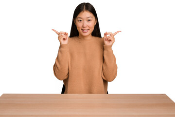 Wall Mural - Young asian woman sitting on a table isolated pointing to different copy spaces, choosing one of them, showing with finger.