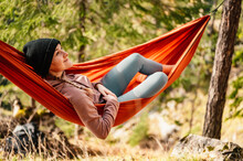 Woman With Cap Resting In Comfortable Hammock During Sunset. Relaxing On Orange Hammock Between Two Trees Pine Enjoying The View
