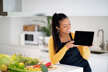 Wall Mural - Happy young african american woman in apron show tablet with empty screen at table with fresh vegetables