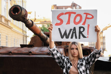 Wall Mural - Sad woman holding poster with words Stop War near broken tank in city