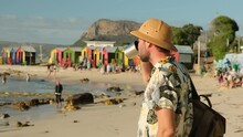A Male Traveler In A Sun Hat Drinks Hot Tea On The Ocean Against The Background Of Colored Houses On The Beach. Hiker In A Shirt With A Backpack Holds A Tourist Metal Mug With Tea And Rests