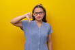 Beautiful Disappointed young Asian woman 30s wearing a dress checkered with glasses showing her thumb down, expressing disapproval isolated on yellow background. people lifestyle concept