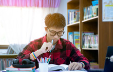 Soft Focus Of Young Asian Boy Wearing Eyeglasses And Rainbow Wristband, Holding Pen, Sitting In Library, Reading Books And Concentrating To Book On Table Before Mid Term Test And Final Test Next Day.