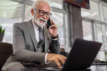 Smiling elderly professional using laptop while talking on smart phone at the office or at train station