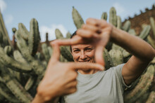 Smiling Woman Looking Through Finger Frame In Front Of Cactus Plants