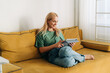 © Ilona - Attractive middle-aged caucasian woman sitting on a sofa works on a computer tablet.