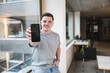 © Westend61 - Smiling young businessman showing smart phone sitting on window sill in office