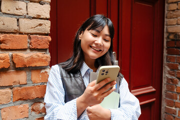 Wall Mural - Young asian girl using mobile phone and holding laptop while standing outdoors