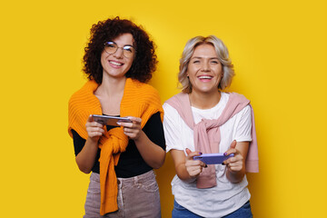 Two smiling young women gamer using smart mobile phone and playing games, isolated over yellow color background. Lifestyle and leisure with hobby concept