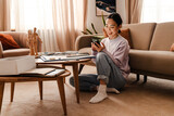 Fototapeta  - Smiling asian woman using mobile phone while sitting on floor at home