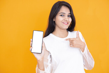 Beautiful young indian girl showing pointing blank display smart phone isolated over orange yellow background, Asian female holding mobile phone with empty screen to put your advertisement.