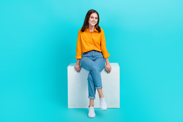 Wall Mural - Full length photo of good mood pretty girl with bob hair dressed yellow shirt sitting on white cube isolated on blue color background