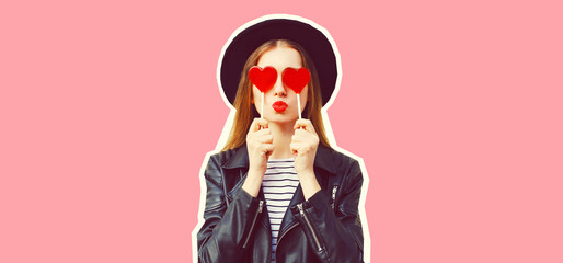 Portrait of stylish young woman with heart shaped lollipop posing wearing black round hat on pink background, magazine style