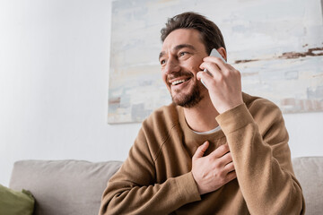 Wall Mural - happy man in beige jumper talking on smartphone at home.