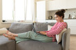 © deniskomarov - A young, cheerful woman spends her time on her laptop lying on the sofa in the comfort of her home, staring at the monitor and texting