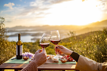 Man and woman cheers red wine glasses at beautiful sunset at outdoor picnic. Romantic dinner at outdoor. 