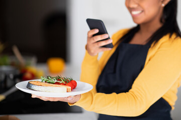 Wall Mural - Millennial african american woman in apron taking photo of fish on phone for blog and social media