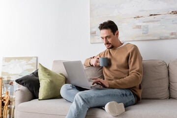 Wall Mural - bearded man using laptop while holding cup and sitting on sofa in living room.