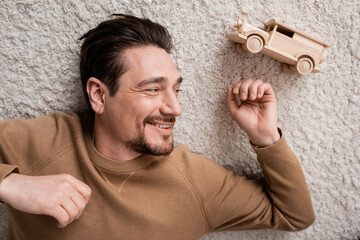 Wall Mural - top view of bearded and cheerful man lying on carpet near wooden toy car in living room.