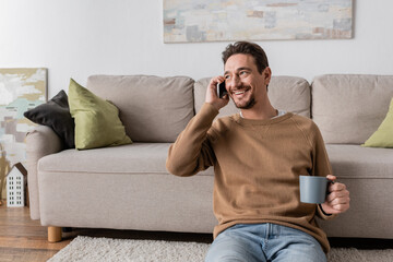 Wall Mural - cheerful man in beige jumper talking on smartphone while sitting with cup of coffee at home.