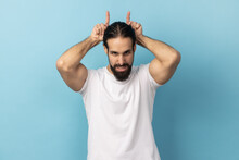 Portrait Of Aggressive Bully Man With Beard Wearing White T-shirt Showing Bull Horns Gesture Over Head, Frowning As Before Attack, Indoor Studio Shot Isolated On Blue Background.