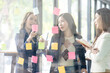 © David - Young creative business Asian woman standing with her colleagues writing new ideas on sticky notes over glass wall, business marketing strategy or User experience ux ui concept.