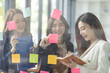 © David - Young creative business Asian woman standing with her colleagues writing new ideas on sticky notes over glass wall, business marketing strategy or User experience ux ui concept.
