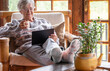 © luciano - Smiling relaxed pensioner woman sitting at home in an armchair using a digital tablet to see videos or reading a book. Elderly smiling caucasian woman in mountain chalet enjoying tech and social media